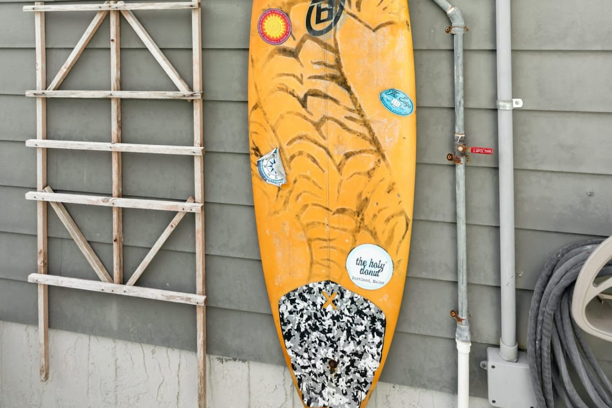 An outdoor shower plumbed to the wall behind a decorative surfboard, with a rack for the boards themselves. Rinse the salt and sand off out here and the floors inside stay very much happier all week long.
