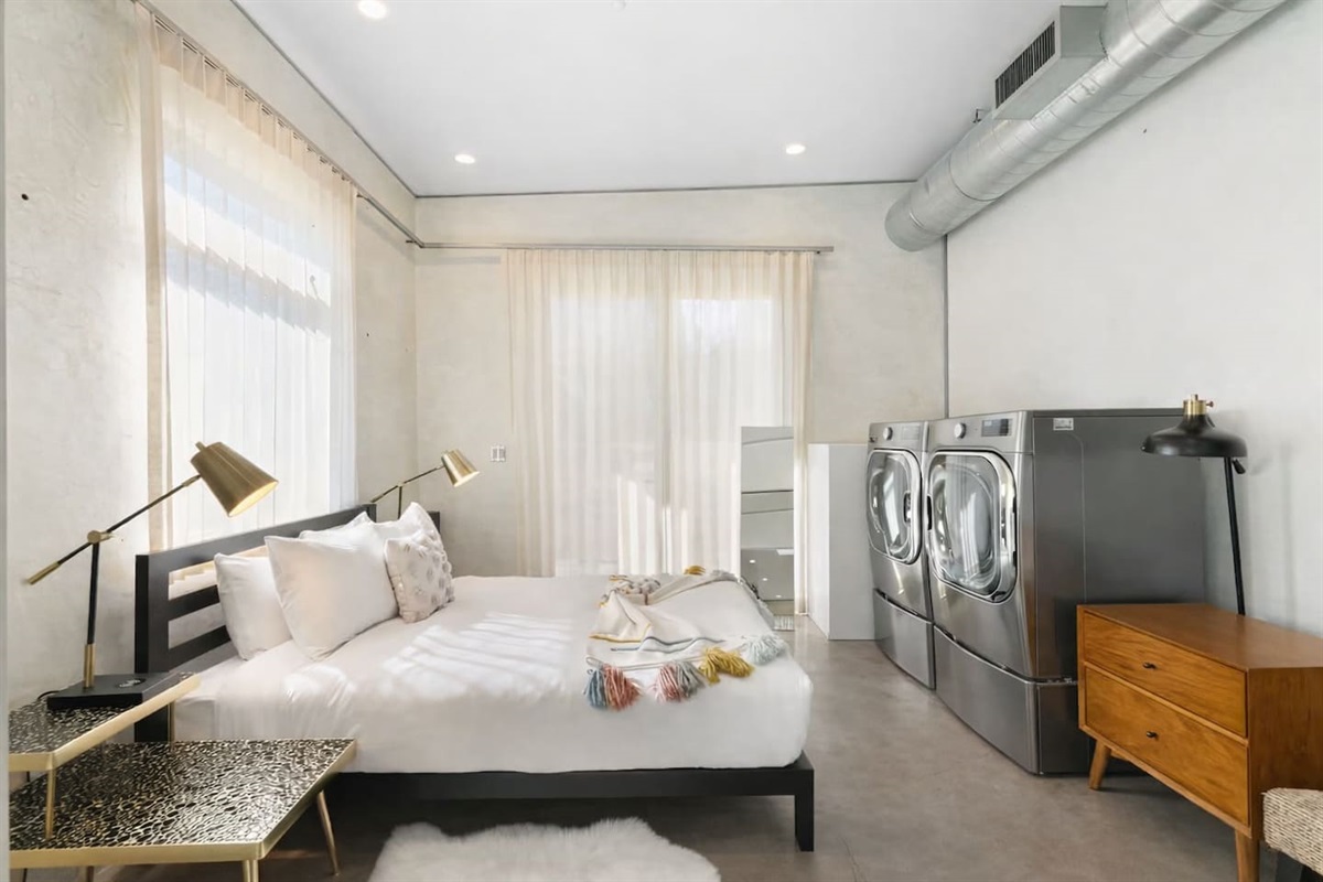 Bright modern bedroom with a cozy bed, floor-to-ceiling sheer curtains, and in-room washer and dryer for added convenience.