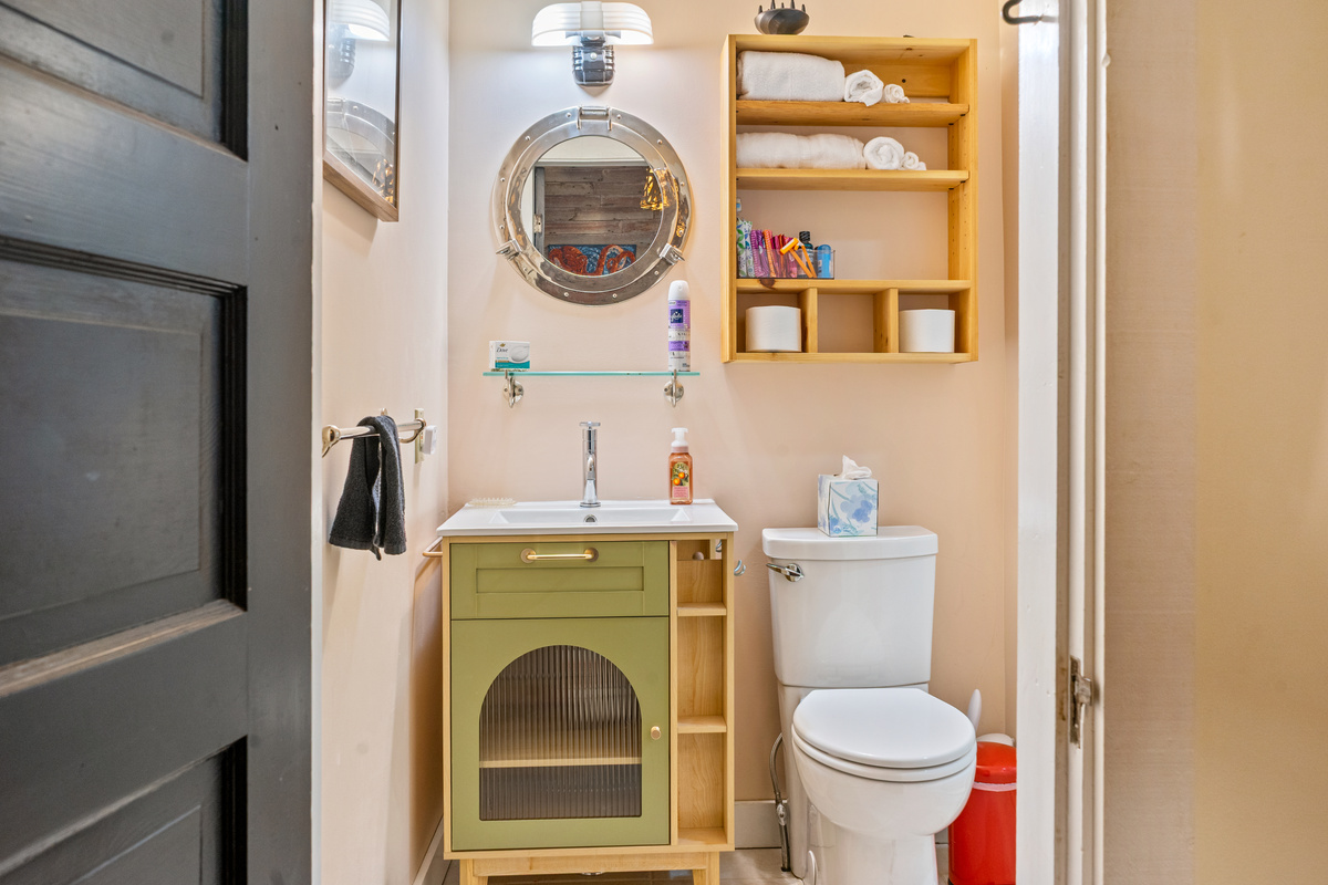 Tucked into the Captain’s hallway, this washroom blends utility with maritime character. A new sage green vanity with a ribbed glass door and a tall chrome faucet sits beneath an authentic porthole mirror, serving the primary wing.