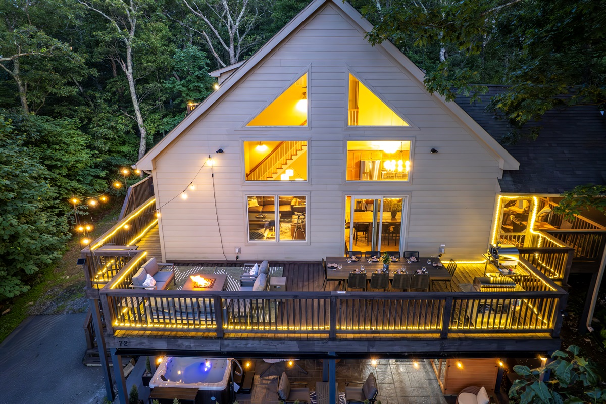 Evening view highlights the fire pit lounge, hot tub, dining zone, and screened game room—all lit with warm café lights and surrounded by forest 