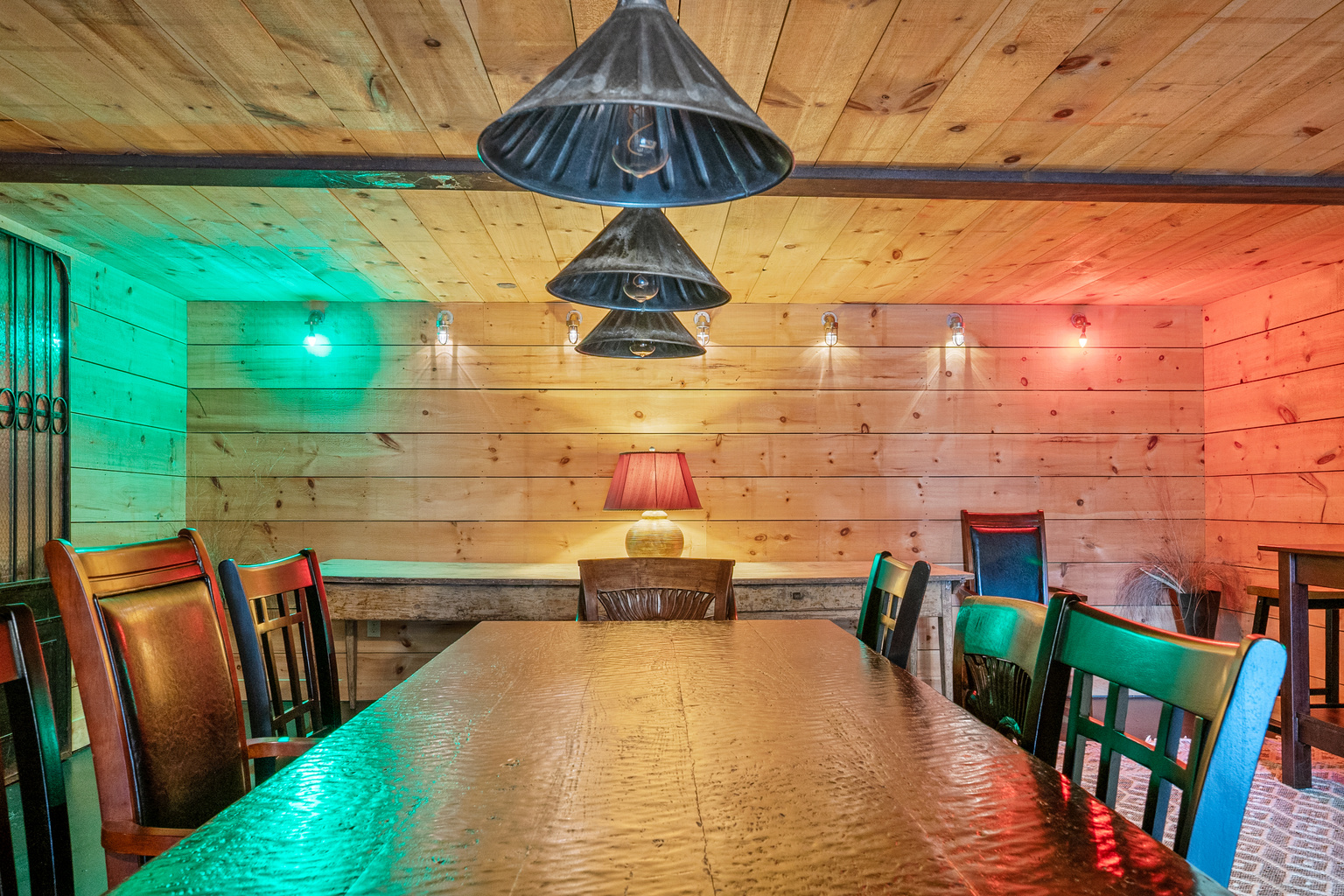 A captain’s view from the head of the mess hall table. Industrial pendant lights align over the hand-scraped wood grain, splitting the space into vivid starboard green and port red zones for an unforgettable, immersive dining experience below deck.