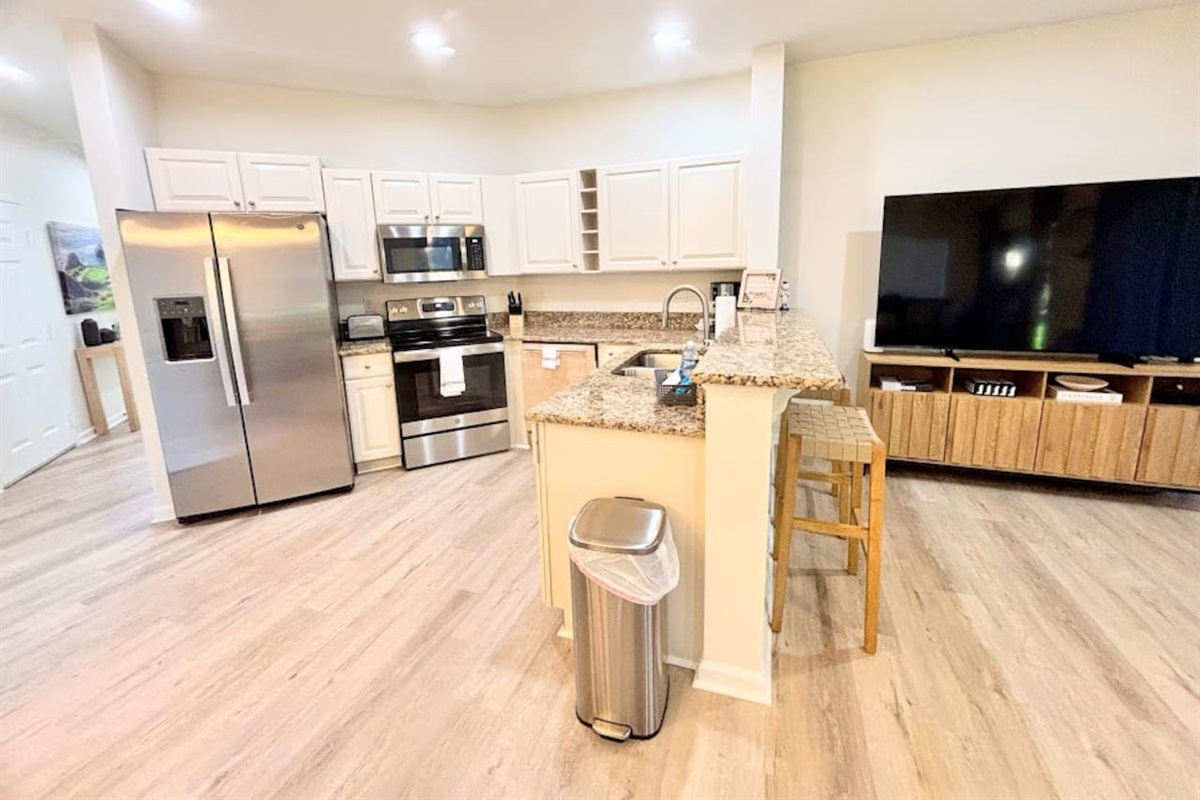 Modern open-concept kitchen and living area featuring stainless steel appliances, granite countertops, breakfast bar seating, wood-look flooring, and a large TV for relaxing or entertaining.