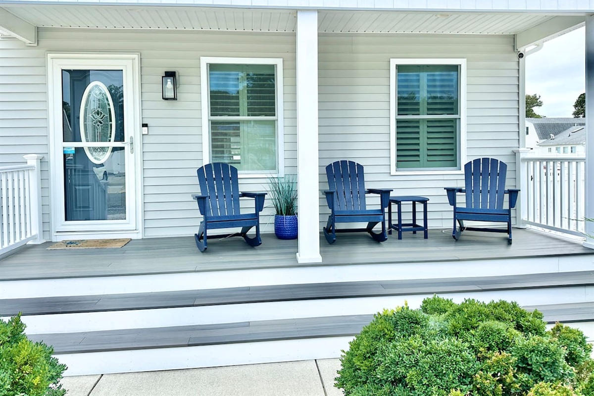 This welcoming front porch features three blue Adirondack rocking chairs and a matching side table, perfect for morning coffee. The grey composite decking and white railings provide a clean, modern look to this charming coastal entryway.