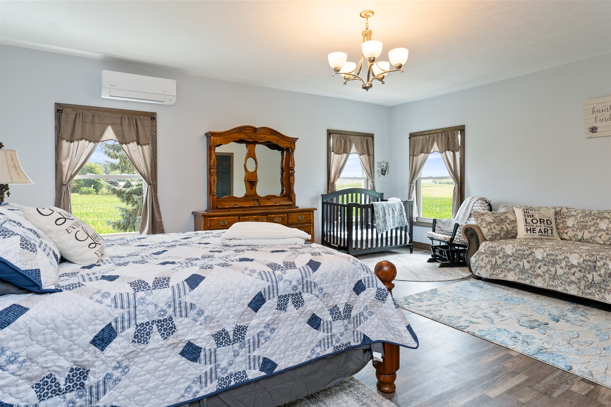 A bright and spacious master bedroom featuring a cozy queen bed, Queen fold out sofa and crib with elegant wood furniture, and stunning countryside views through large windows. Perfect blend of comfort and charm for a restful stay.