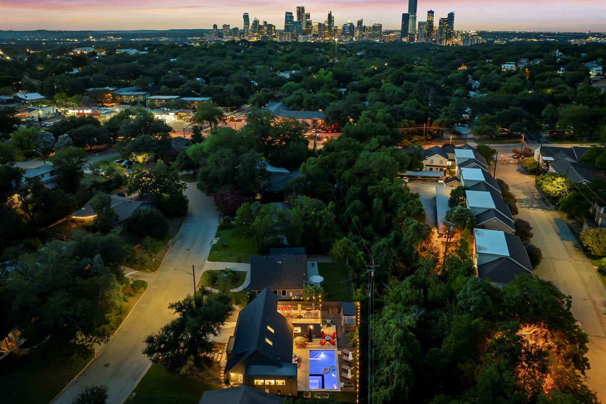 An aerial view of a modern home with a swimming pool and illuminated deck, nestled in a lush, tree-filled neighborhood with the vibrant city skyline visible in the distance.