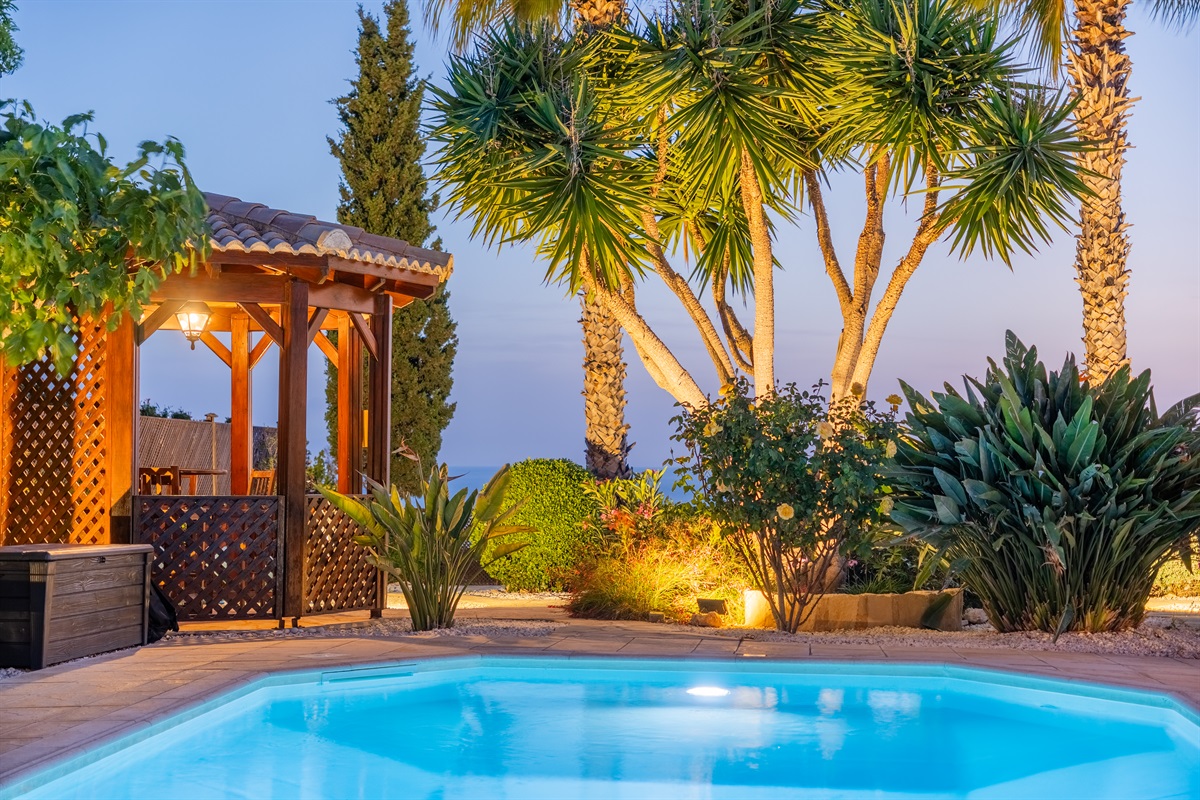 As the sun sets, the garden transforms into a magical retreat—spotlights accentuate the trees, while string lights in the gazebo and outdoor kitchen create a warm, inviting ambiance.