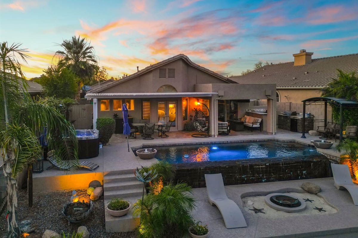 Every corner of this backyard has something to offer — hot tub, pool, fire pit, outdoor kitchen, and a covered lounge — all bathed in the warm glow of a stunning desert sunset.