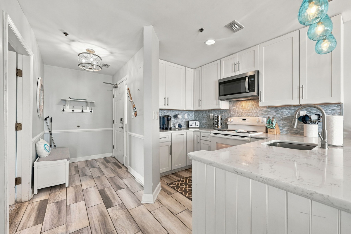 Welcoming entryway hall transitioning into a modern coastal kitchen featuring light wood-plank flooring, white shaker cabinetry, shimmering mosaic tile backsplash, and a breakfast bar island with light granite countertops.   