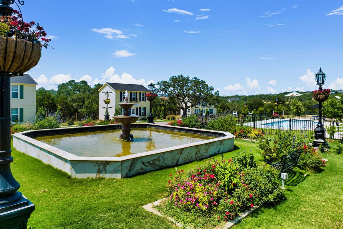 Stroll through manicured gardens centered around a classic fountain, with rolling hills and blue skies as your backdrop.