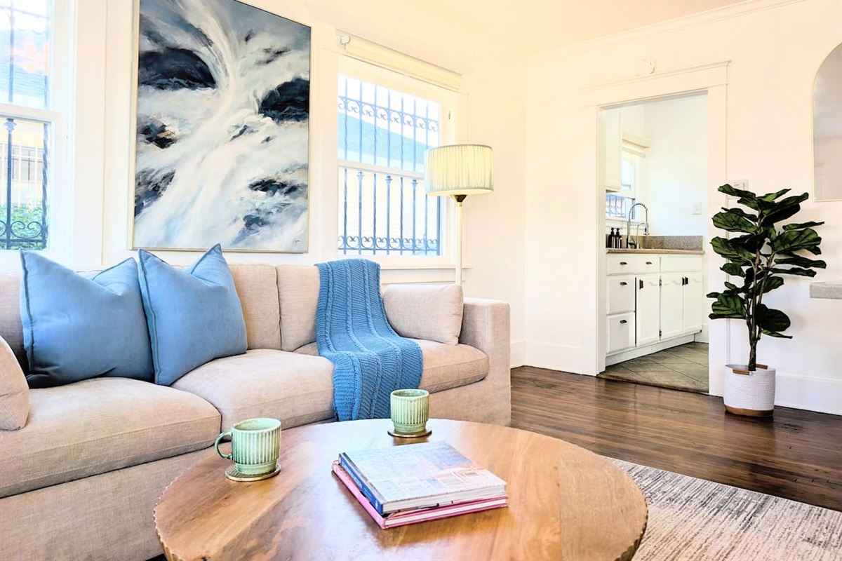 This bright, cozy living roomradiates an airy, coastal charm. Natural light floods the space, illuminating the warm hardwood floors and a plush, inviting beige sofa layered with vibrant blue accents. Framed by clean white walls, an abstract ocean painting 