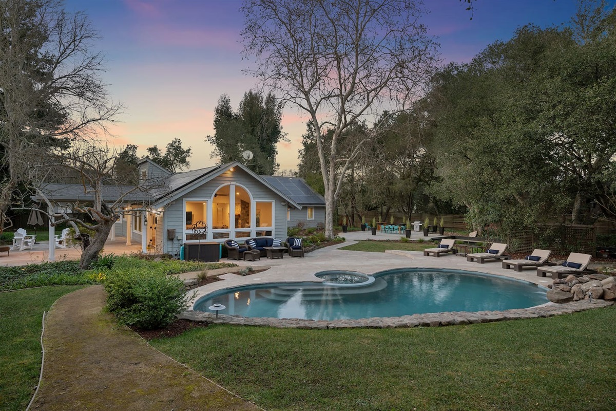 To the left, a pool table and seating area outside the kitchen and living room doors. In front, a pool, spa, rock waterfall, pool loungers, and couch + accent chair relaxation space. To the right, the dining pavilion and bocce court.