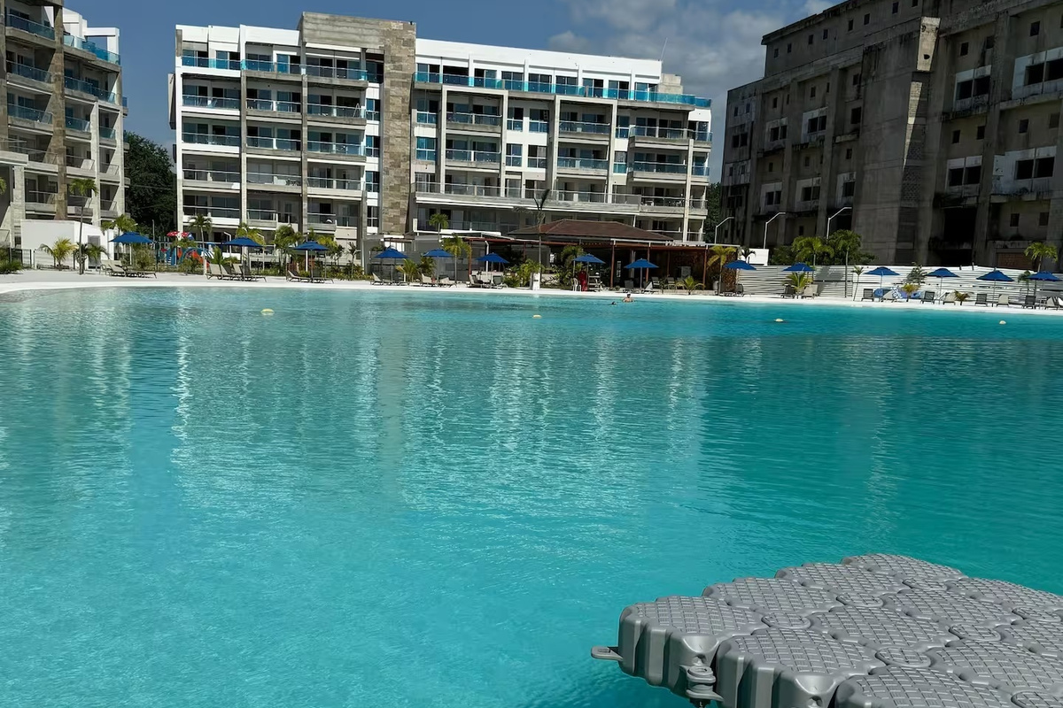 Gran piscina tipo laguna con tranquilas aguas turquesas, asientos junto a la piscina y acceso a un muelle flotante como parte de las amenidades compartidas del resort.