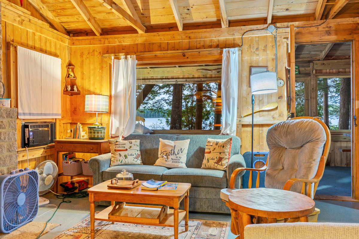 Cozy living room at Loonrise Point looking toward the lake! Features a plush sofa, glider chair, window AC unit, and supplementary fans for warm summer days. Door on right opens directly onto the screened porch on Beech Hill Pond in Otis, ME.