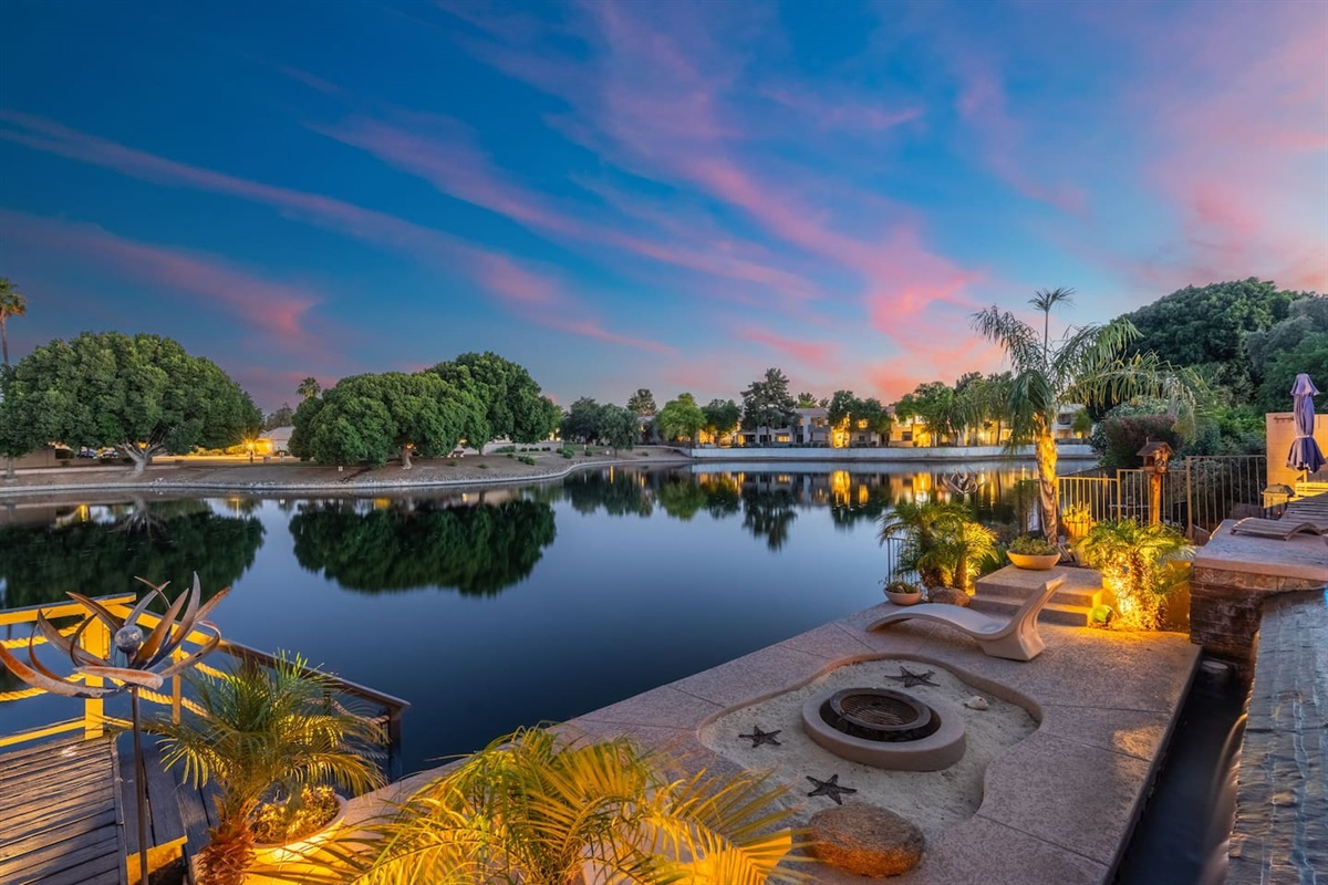 The lakeside fire pit glows under a breathtaking sunset sky — gather here after dark to relax, connect, and end the day in the most beautiful way possible.
