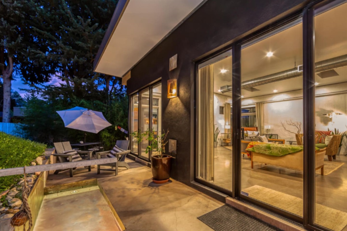 Inviting twilight view with floor-to-ceiling glass doors, warm interior lighting, and relaxed outdoor seating under a large umbrella.