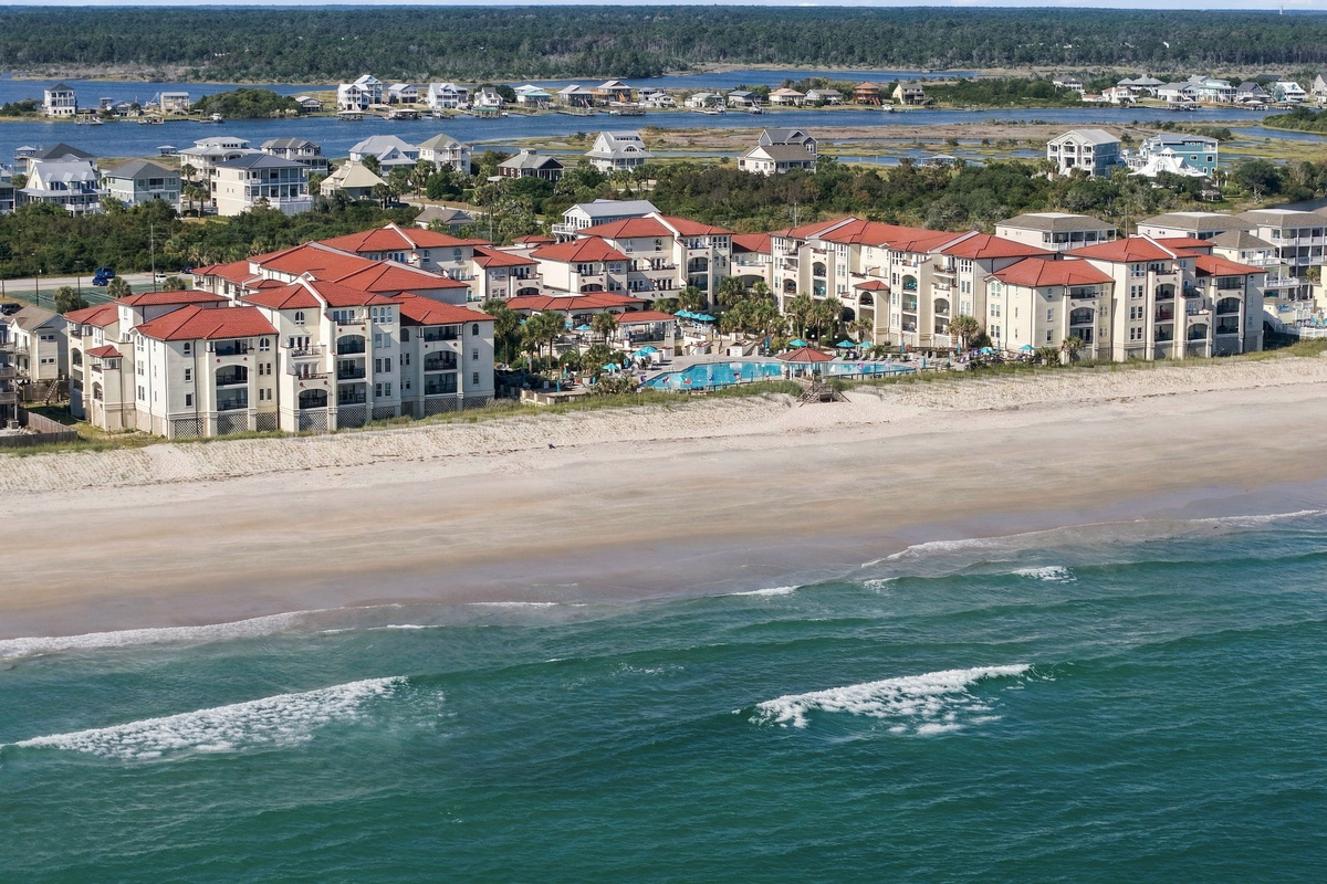 Coastal aerial perspective showing the complex situated directly along the shoreline, featuring beachfront buildings, pool amenities, and water views on both sides.