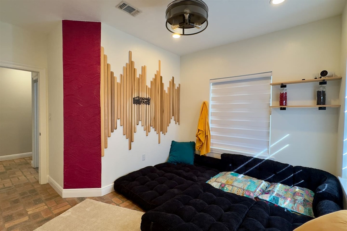 Stylish lounge area with a plush black sectional, artistic wood wall feature, and warm accent tones. Natural light, ceiling fan, and cozy decor create a relaxing retreat.
