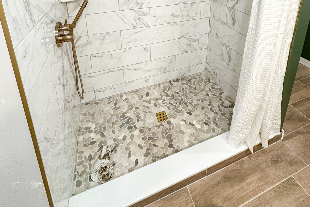 Custom tile walk-in shower featuring a rainfall shower head, handheld wand, pebble stone floor, and warm brass fixtures. A spa like space designed to help you relax after a day at the beach, pool, or local wineries. (Glass doors coming in mid July)