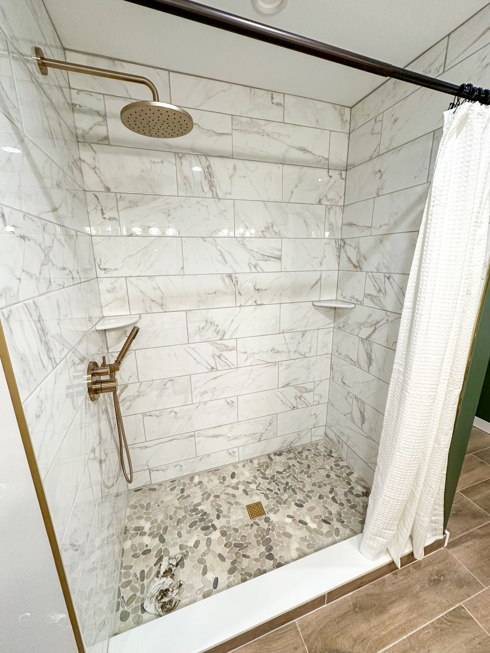 Custom tile walk-in shower featuring a rainfall shower head, handheld wand, pebble stone floor, and warm brass fixtures. A spa like space designed to help you relax after a day at the beach, pool, or local wineries. (Glass doors coming in mid July)