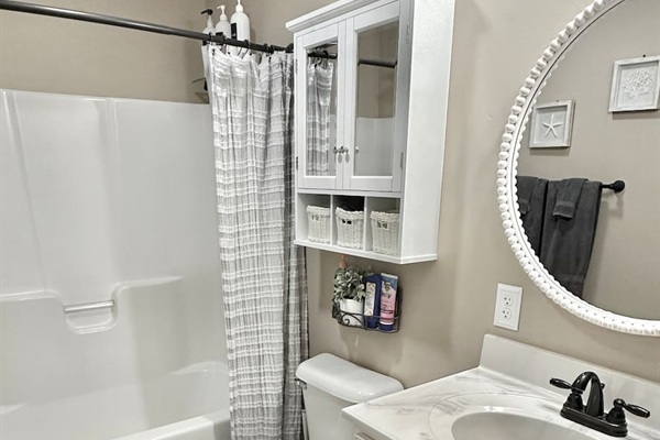 This bathroom includes a white bathtub and shower combo with a textured grey curtain. A white wall cabinet with glass doors and storage baskets hangs above the toilet, while a large beaded round mirror sits above the vanity.