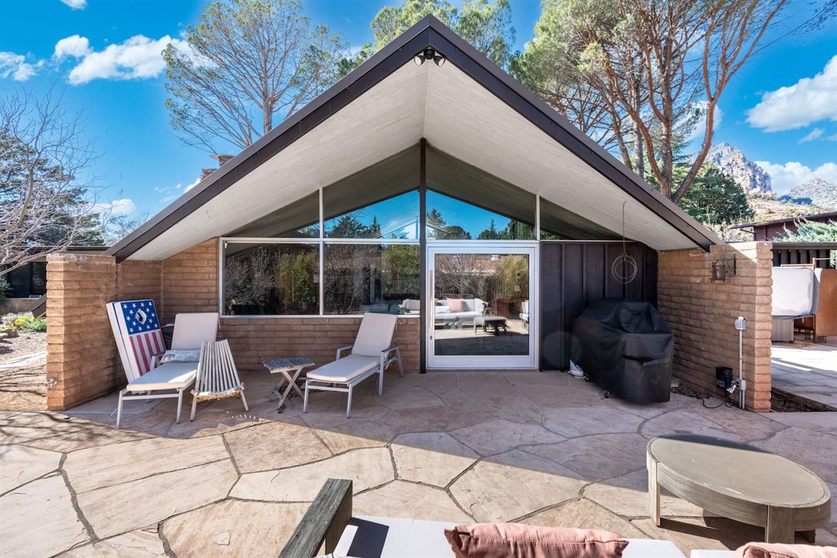 Striking mid-century architecture with dramatic vaulted rooflines and floor-to-ceiling glass, opening onto a private patio with lounge seating and grill — a perfect blend of indoor-outdoor living in a stunning desert setting.