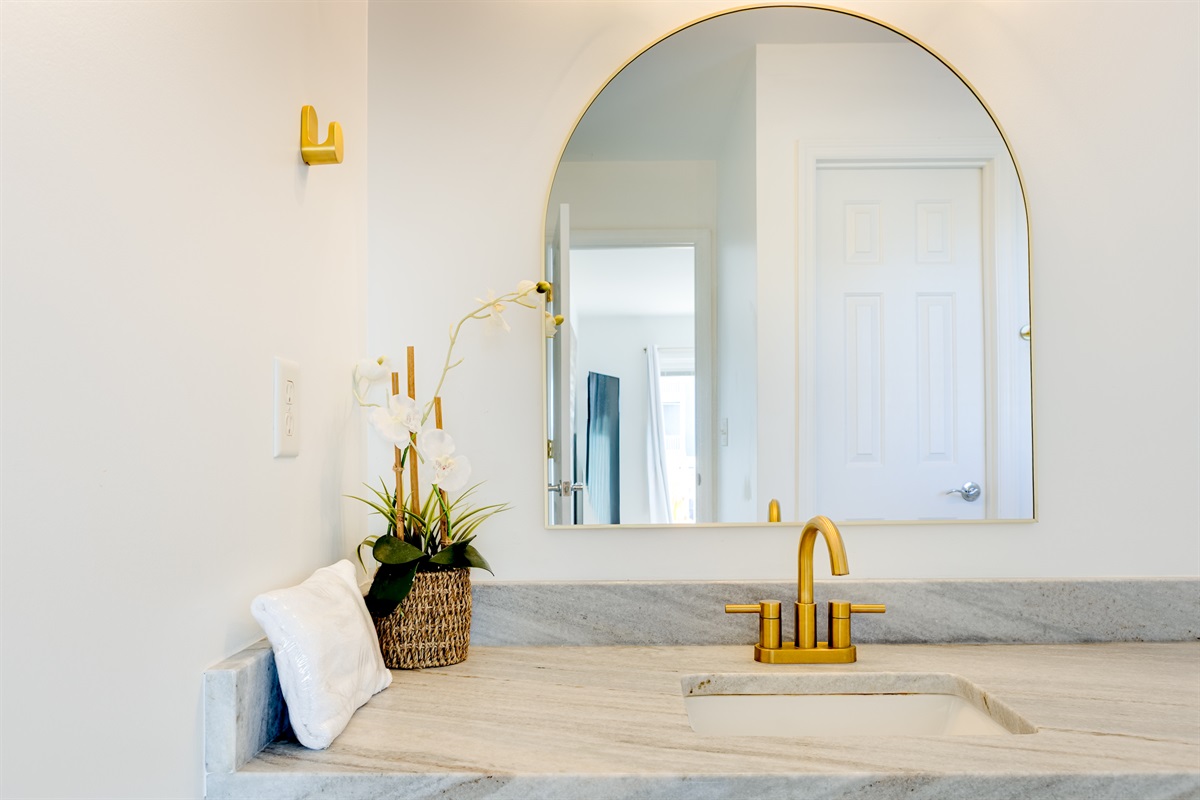 Bright and modern vanity with a marble countertop, elegant gold fixtures, and a large arched mirror