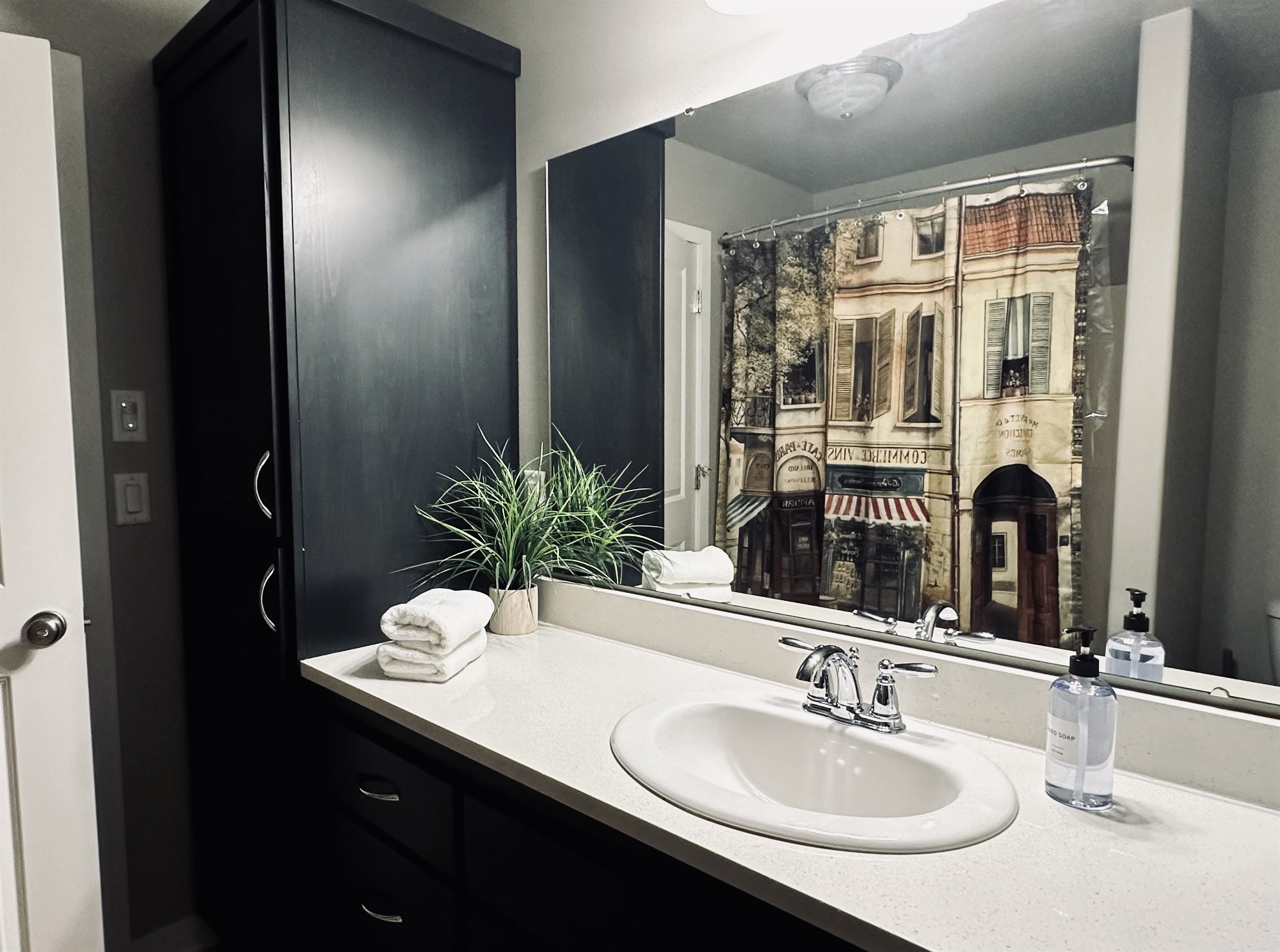 A modern bathroom features a white countertop with a sink, neatly stacked white towels, a small potted plant, and a bottle of soap, with a large mirror reflecting a vintage-style shower curtain depicting a quaint street scene.
