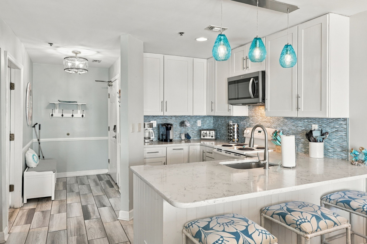 Modern kitchen and entry foyer featuring a breakfast bar with cushioned counter stools, vibrant aqua glass pendants, white shaker cabinets, and glass mosaic backsplash tile.