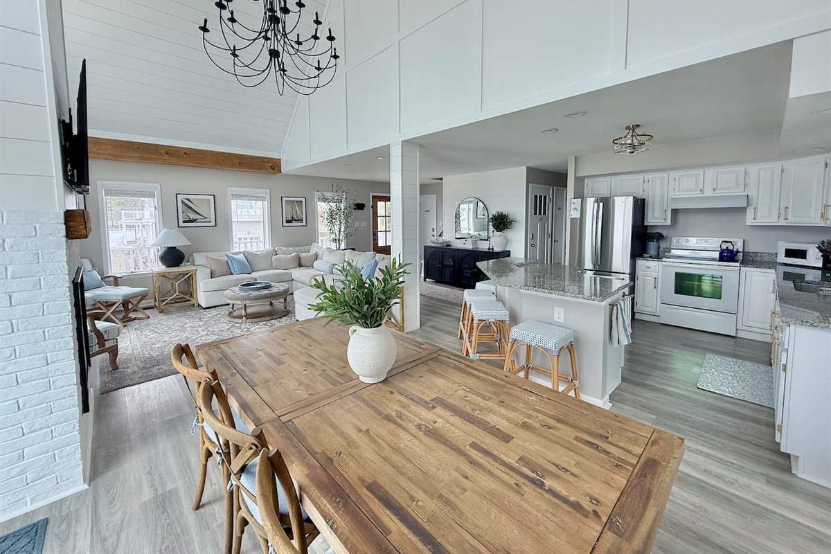 Enjoy an open-concept living space highlights a dining area with a wood table, a cozy lounge with a sofa, and a modern kitchen. A vaulted ceiling and chandelier create an airy, inviting atmosphere throughout the home.