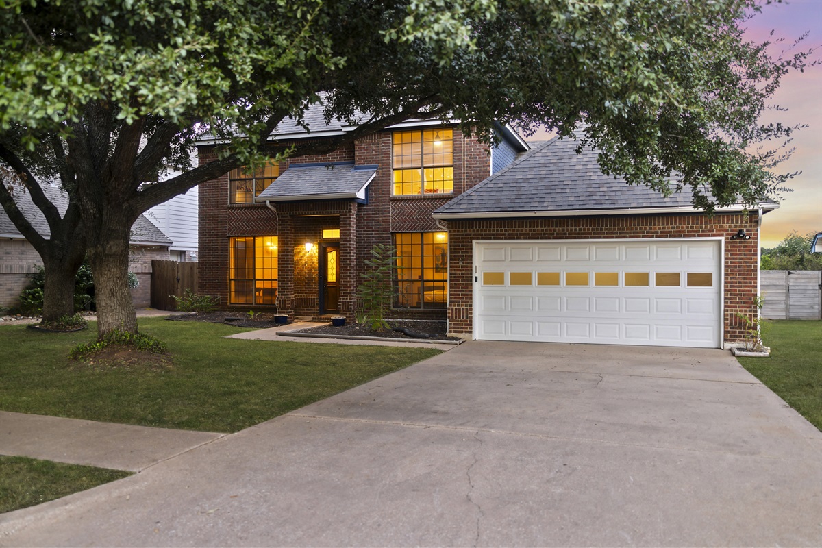 Beautiful two-story brick home with a spacious driveway, large front yard, and mature trees. The warm lighting from the windows invites you into a cozy, welcoming space perfect for families or group stays.