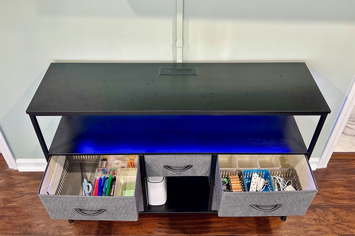 Living room TV console with deep storage drawers stocked for everyday convenience. Includes office and household essentials such as notepads, pens, scissors, batteries, charging cables, and an extension cord.