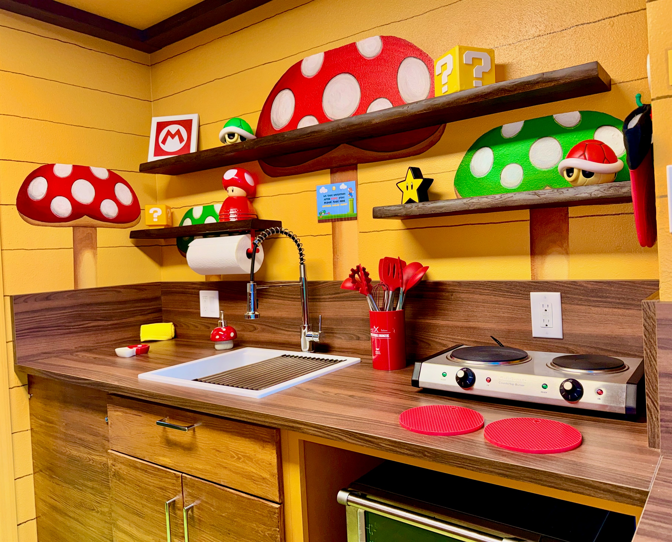 Fully equipped Nintendo-inspired kitchen! Warm wood cabinets, dark countertops, built-in microwave, sink, and vibrant Mario mushroom shelves stocked with fun details—perfect for family meals at Mario's Mini Mansion.