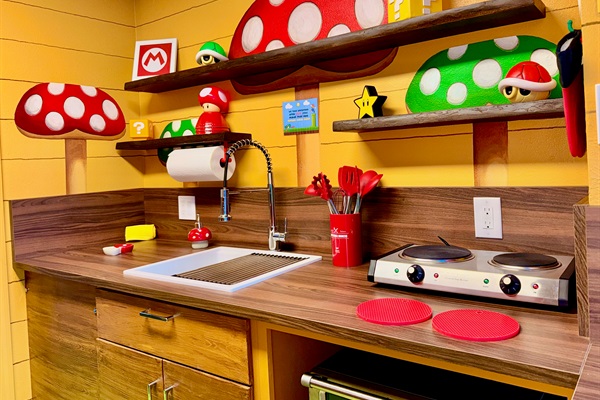 Fully equipped Nintendo-inspired kitchen! Warm wood cabinets, dark countertops, built-in microwave, sink, and vibrant Mario mushroom shelves stocked with fun details—perfect for family meals at Mario's Mini Mansion.