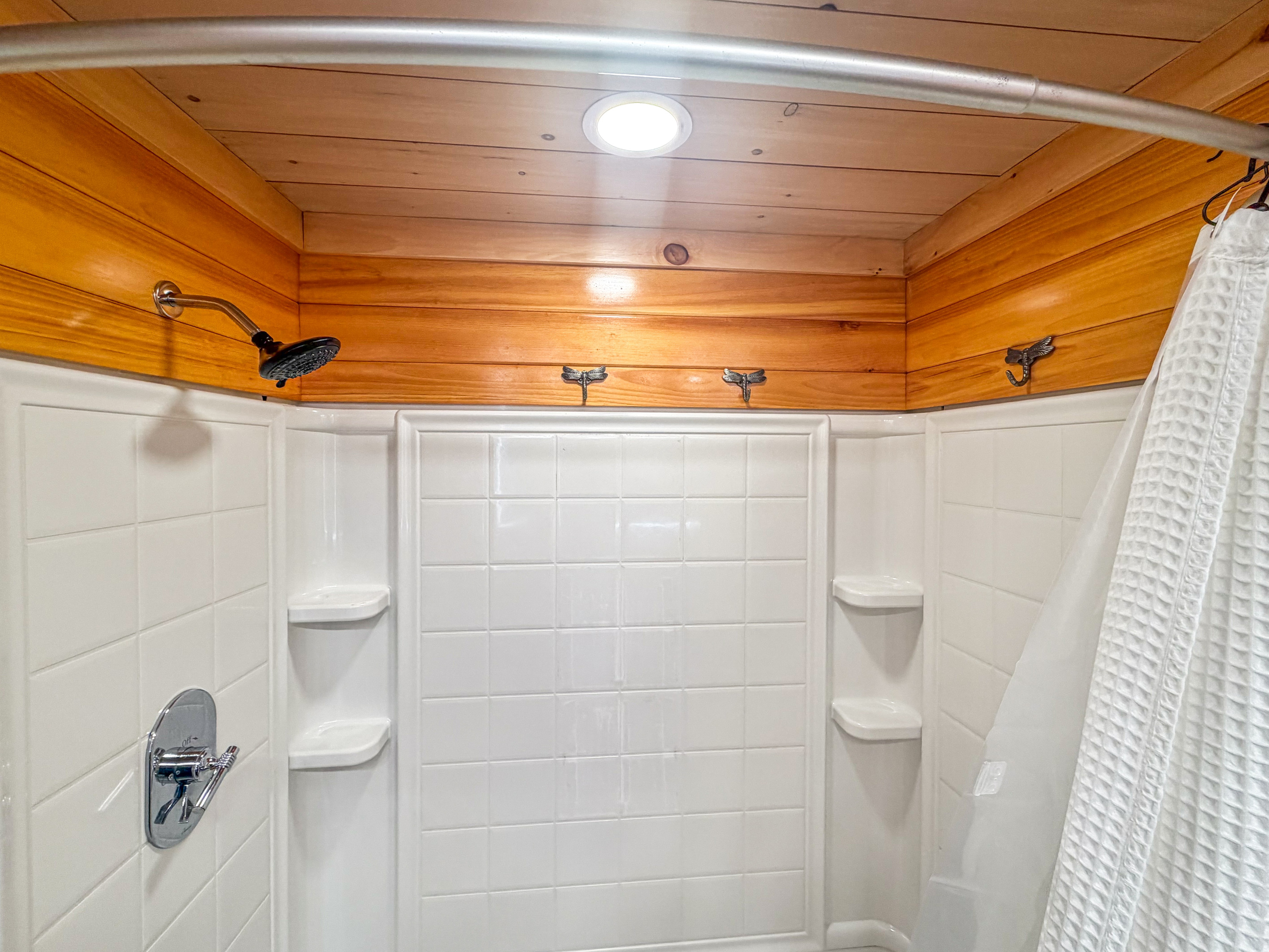 Rinse off comfortably in this pristine white surround shower enclosure. The space is equipped with multiple built-in corner shelves for soaps, decorative dragonfly wall hooks for hanging wet towels, and a dedicated ceiling canned light fixture.
