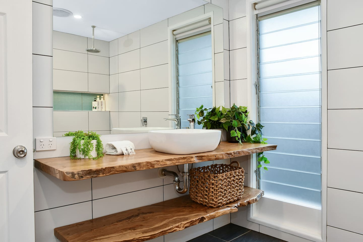 The thick natural-edge timber vanity creates a standout feature in this bathroom at Aaron's Beachside Boat House, paired with crisp white finishes and greenery.