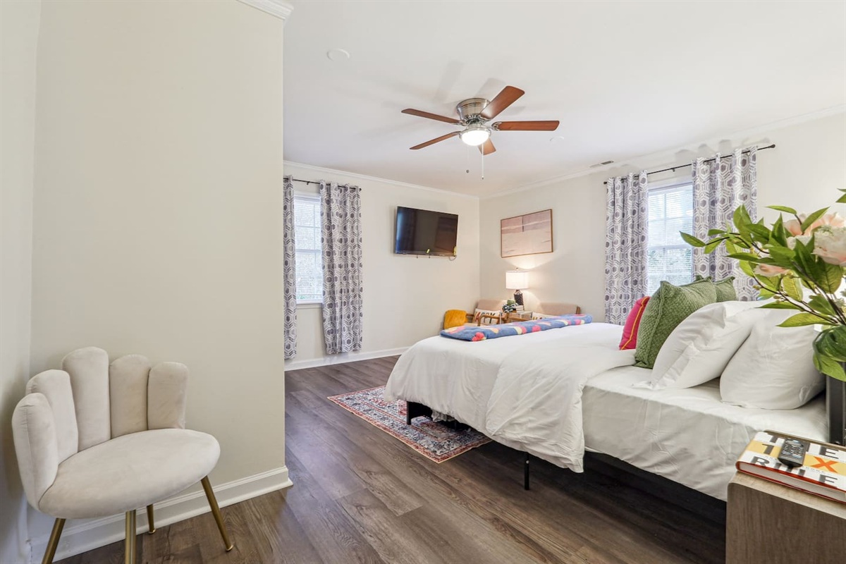 A cozy bedroom featuring a King bed, a ceiling fan, two windows, two chairs, a bedside shelf, a bedside lamp, and a TV — a comfortable space to relax and unwind.
