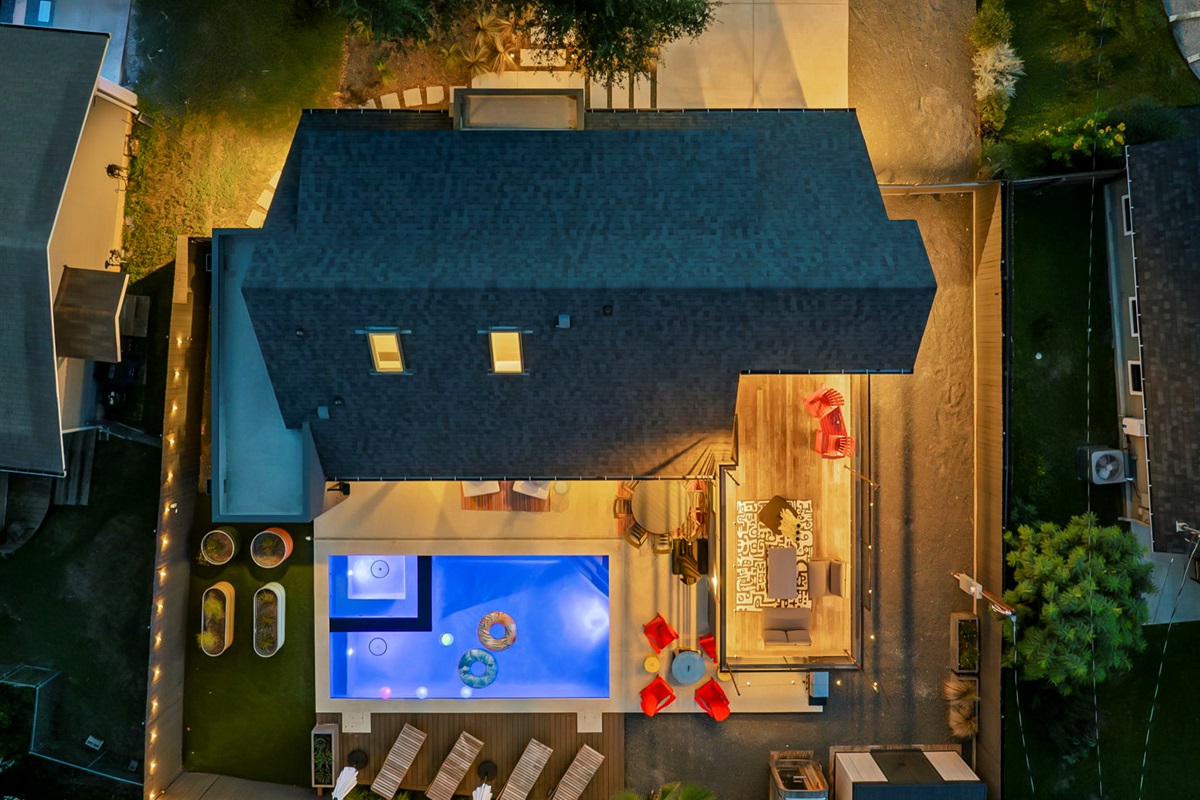 An aerial nighttime view of a modern backyard oasis, featuring a lit swimming pool with a hot tub, a spacious deck with lounge chairs and seating areas, and a lush green lawn, all illuminated to create a perfect space for outdoor entertaining.
