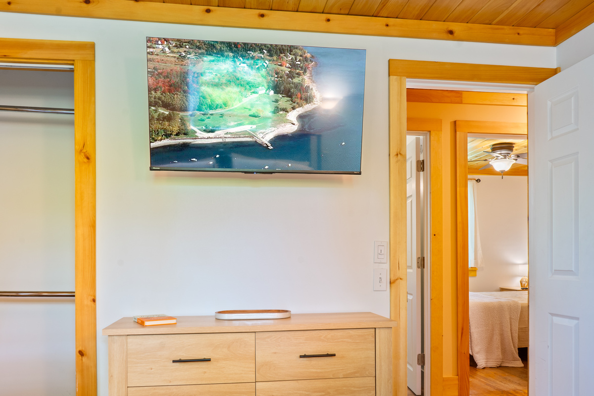 Catch the game on the large wall-mounted flat-screen TV centered above the light-wood dresser. This straight-on view illustrates the layout flow from the primary bedroom suite past the open closet area and directly out toward the main hallway.