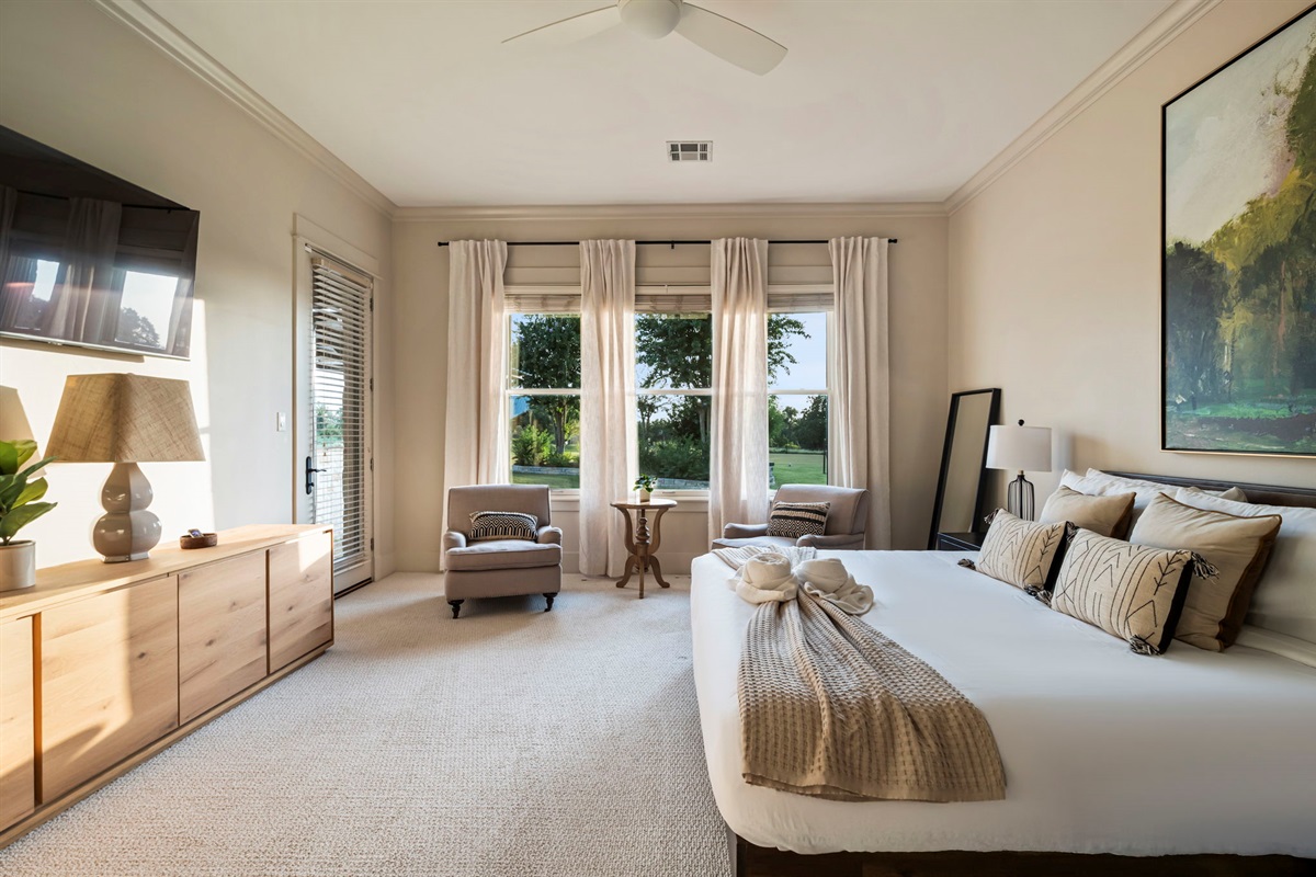 The room includes two cozy lounge chairs with a center table, a full-length mirror, two bedside lamps, and large windows that fill the space with natural light.
