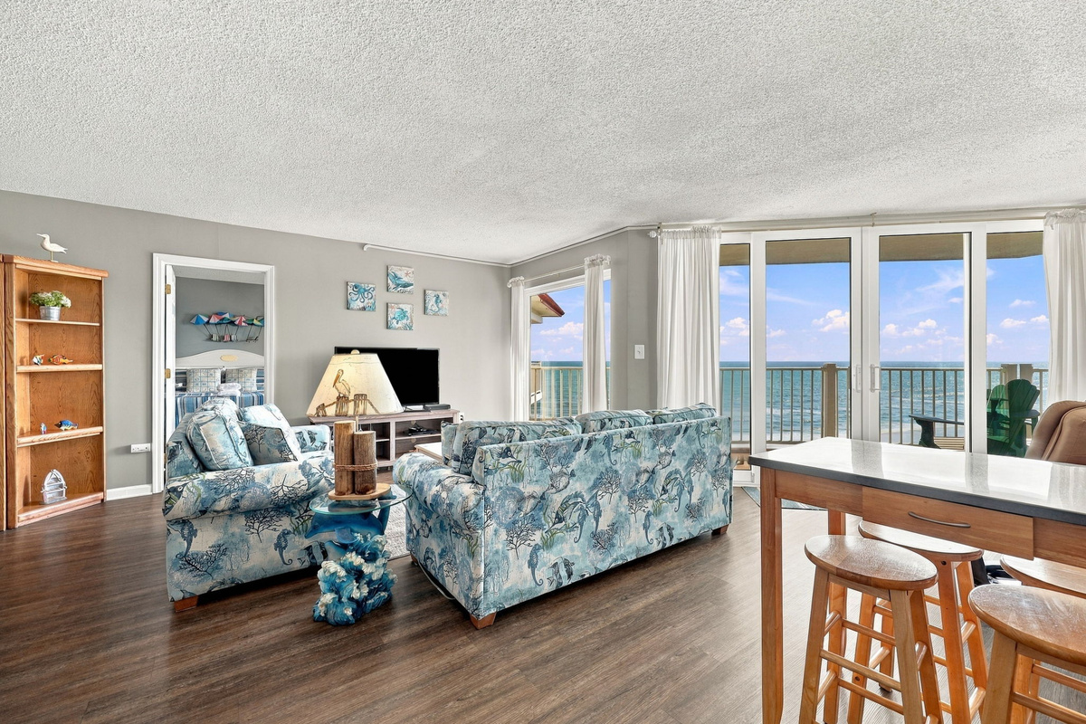 Spacious living room area featuring floral blue seating, wooden accents, and sliding glass doors that open to a beachfront balcony view.