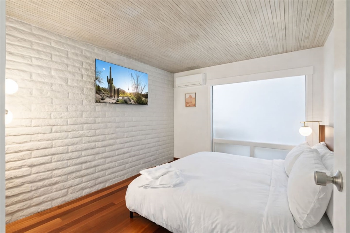 Cozy, modern bedroom with a plush bed, white brick accent wall, warm wood floors, and wall-mounted TV.