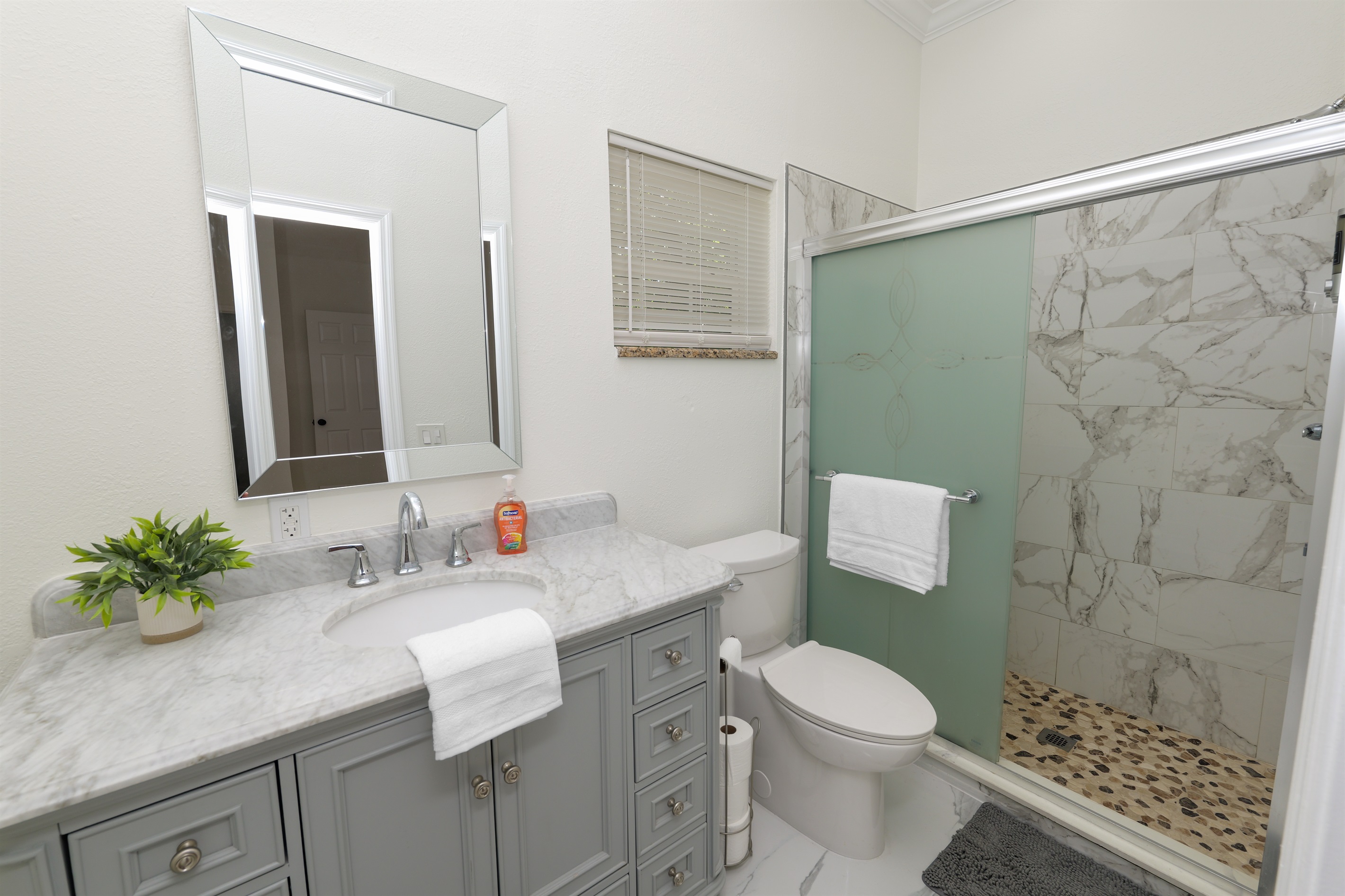 Modern elegance meets function in this beautifully updated bathroom, featuring a sleek marble vanity, stylish gray cabinetry, and a spa-inspired walk-in shower with frosted glass and pebble stone flooring.
