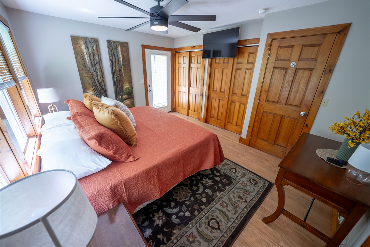 Bright primary suite with a king bed, large windows overlooking the creek, direct access to the deck, Smart TV, ceiling fan, spacious closets, and an attached en suite.