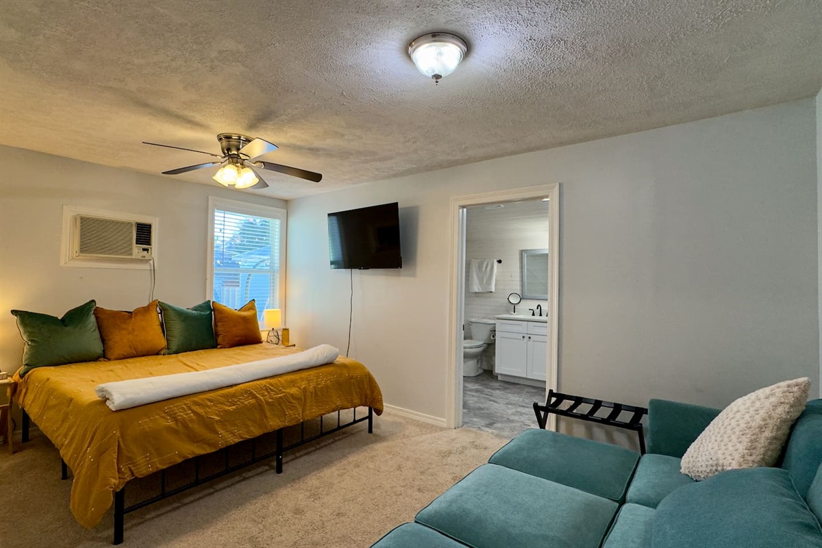 A spacious, comfortable bedroom featuring a plush bed with bold accent pillows, a cozy seating area, wall-mounted TV, and easy access to a private bathroom.