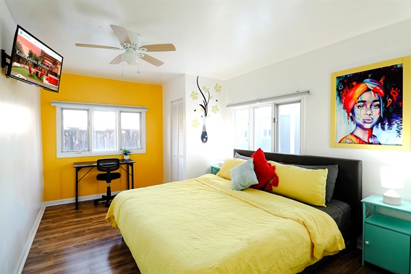 A bedroom with a refreshing mix of yellow and white walls, enhanced with a TV and a ceiling fan, combining visual warmth with airy circulation for a comfortable retreat.