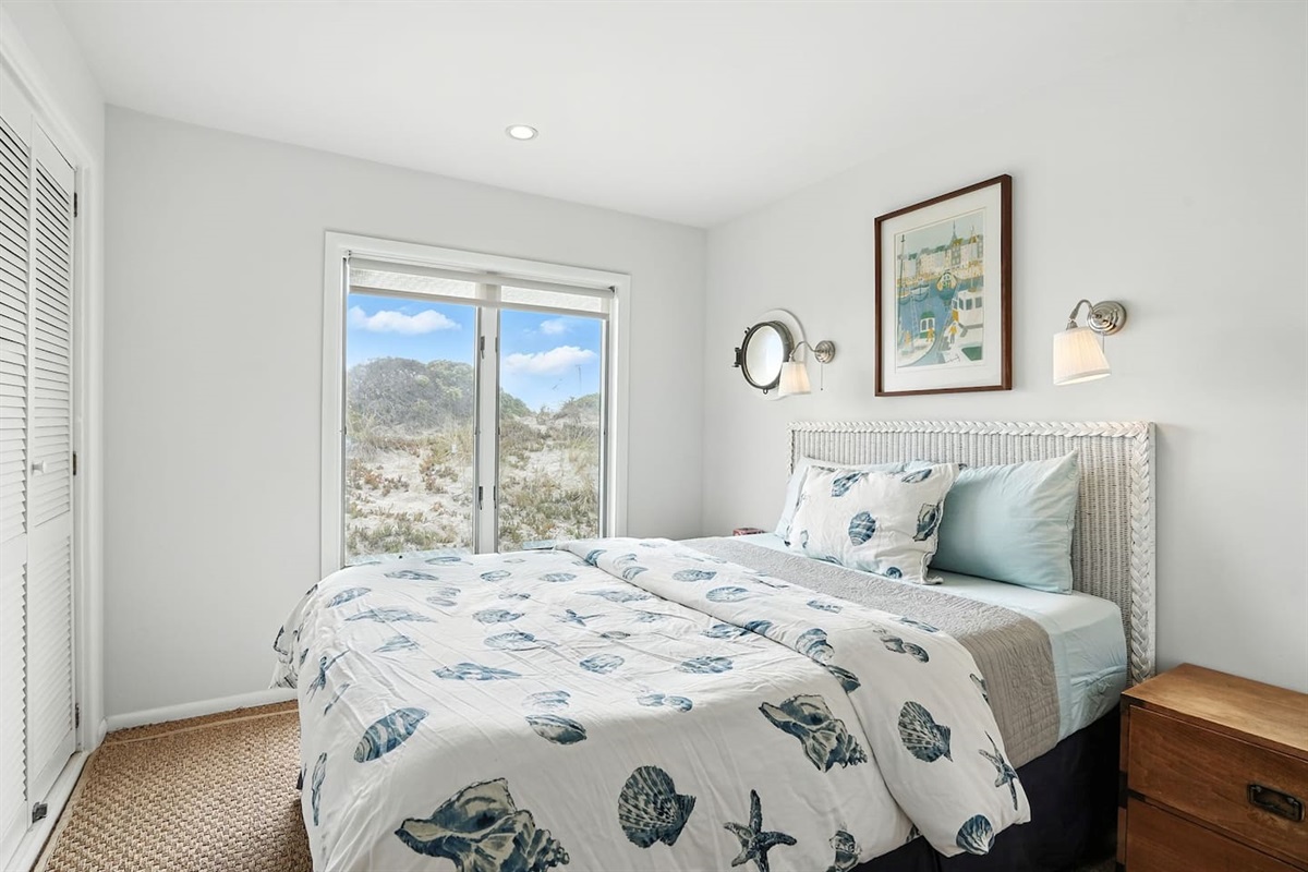 - Light filled bedroom with coastal bedding and sliding glass door to outdoors  
- Features porthole style window, nightstands, and wall mounted lights  
- Serene setting with soft tones and dune views from the bed