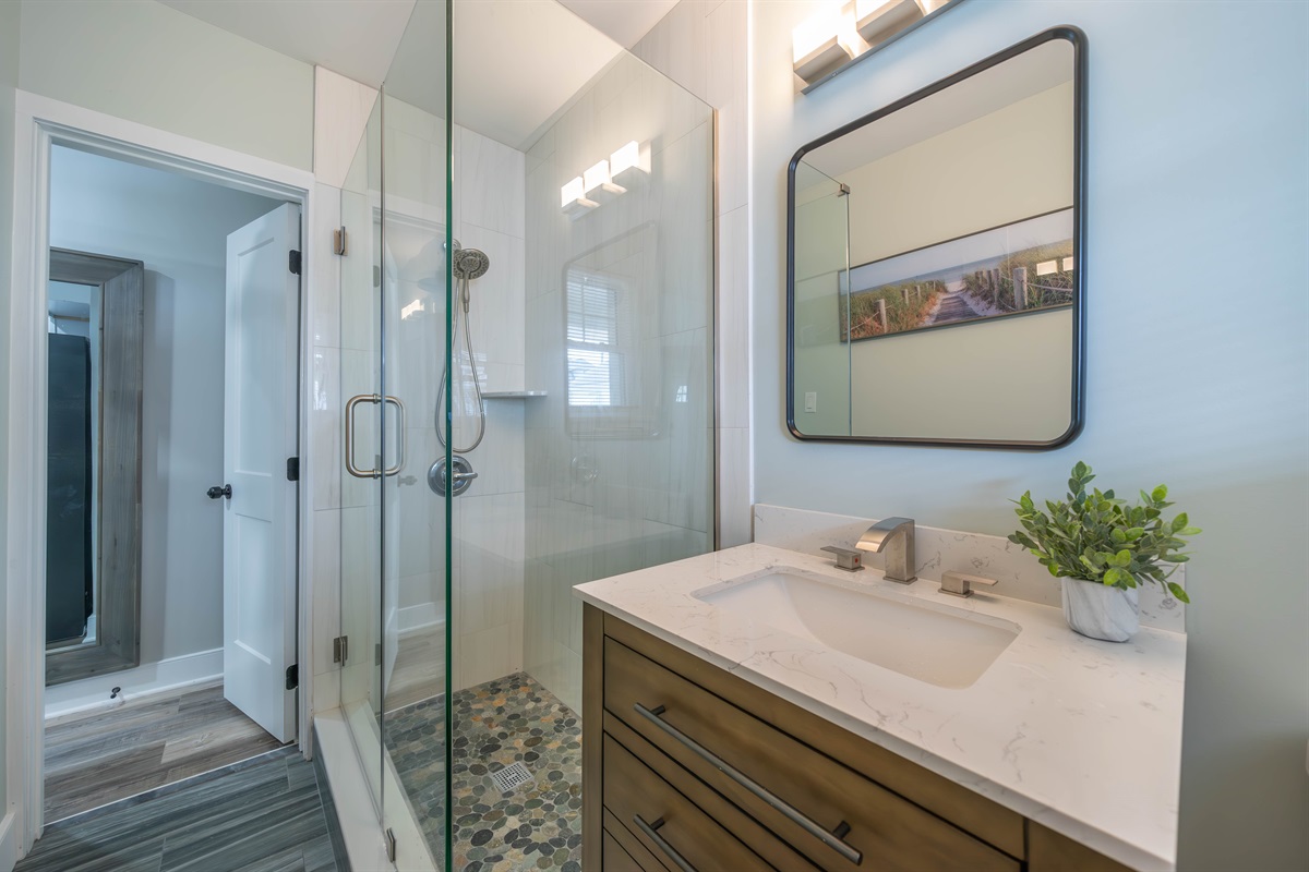 Clean, modern bathroom with a glass walk-in shower, pebble-style tile floor, and bright lighting. The vanity offers generous counter space and a large mirror, creating a fresh and comfortable setting for daily routines.