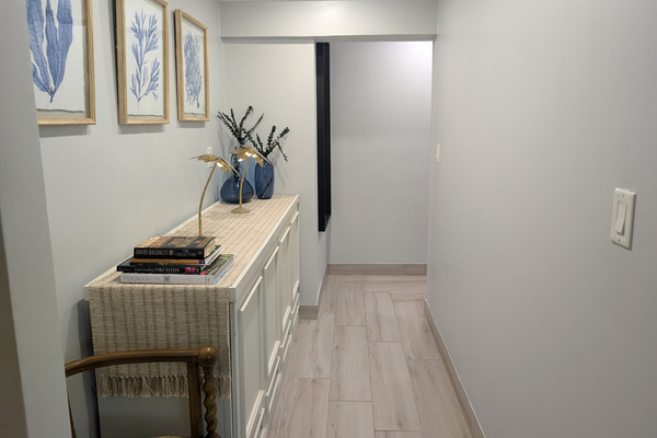 Downstairs hallway at Villa Coral featuring light wood-look tile floors, a white credenza with coastal decor books, blue glass vases, and framed botanical seaweed artwork in Dorado, Puerto Rico.
