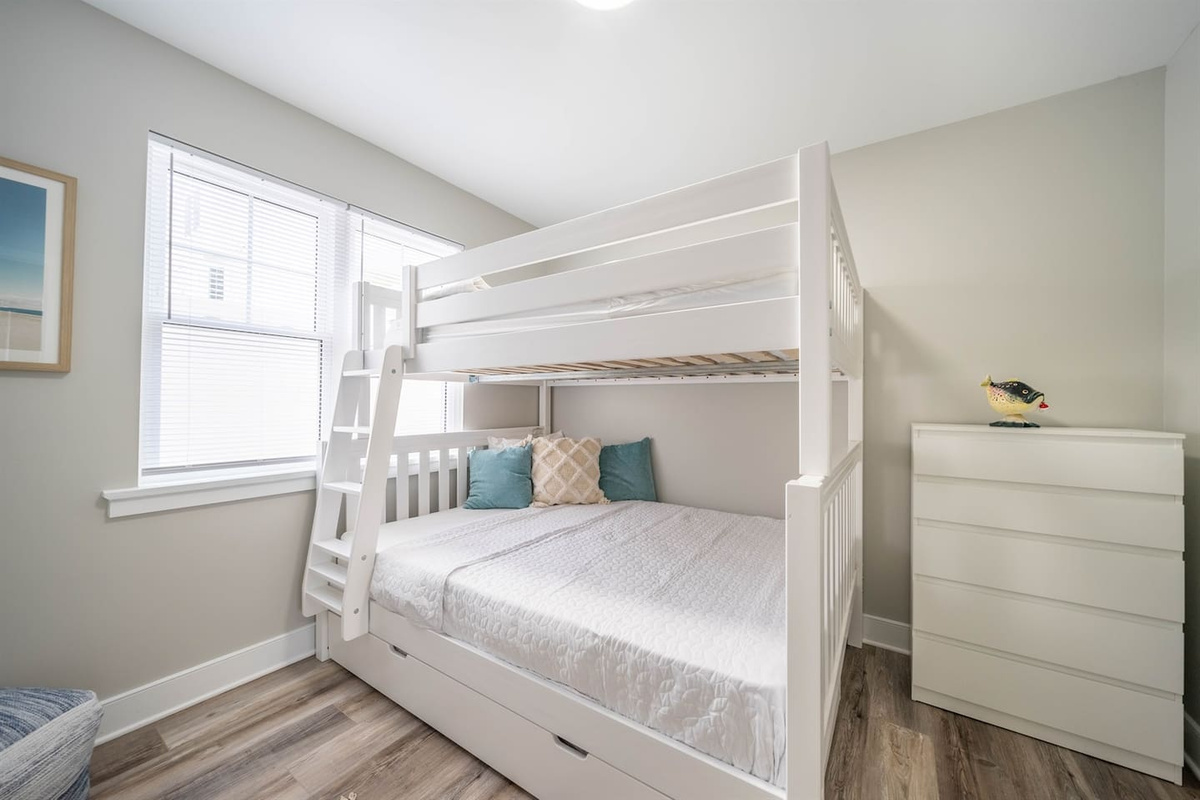 Bright bedroom featuring a white bunk bed with a full-size lower bed and upper bunk. Soft natural light, neutral tones, and simple furnishings create a comfortable space suited for families or groups, with storage drawers built into the bed frame.