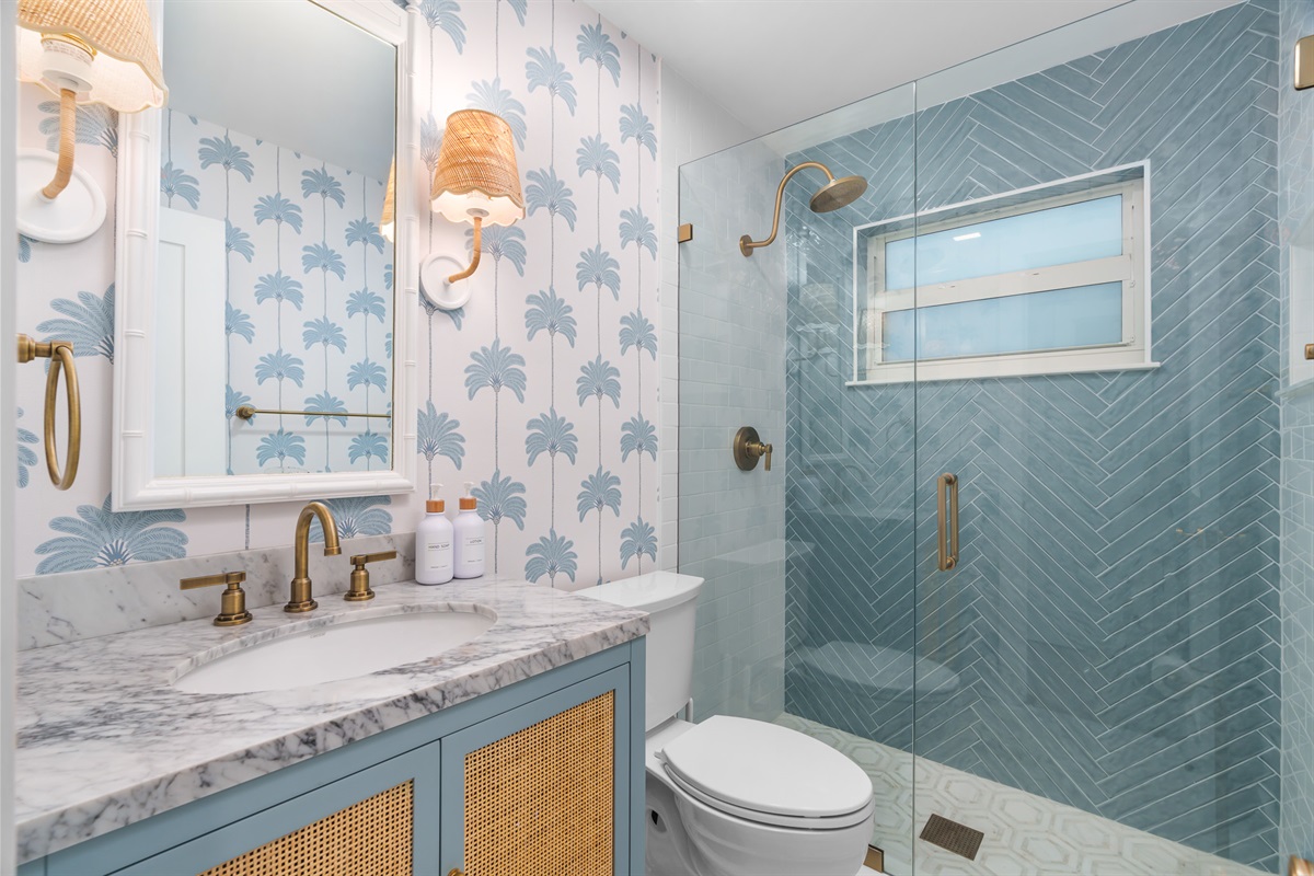 Refresh in this beautifully designed bathroom featuring palm-print wallpaper, marble vanity, brass fixtures, and a spacious glass walk-in shower.